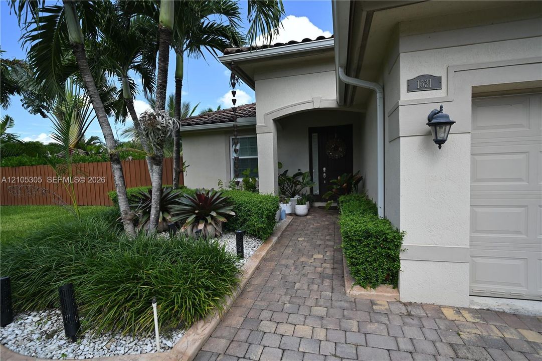 Paver driveway and walkway, arched entryway, exterior wall sconce, and mature landscaping