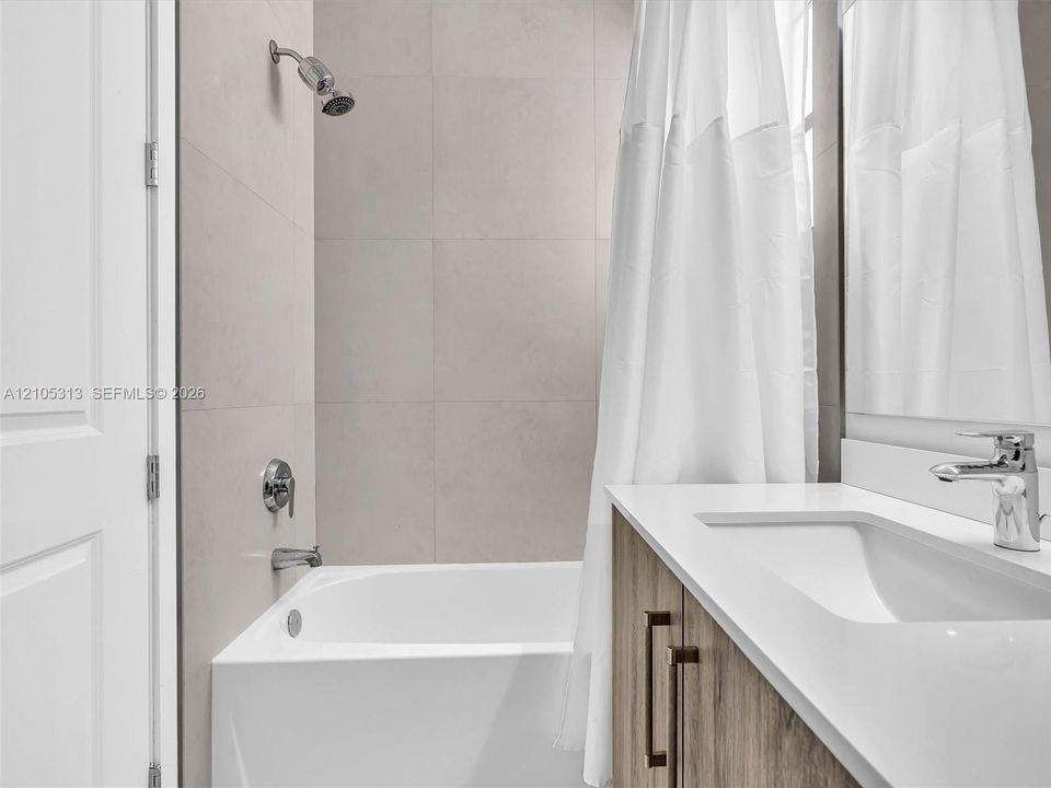 Bathroom featuring a white composite tub with a shower head, full-height neutral wall tile, wood-finish vanity with a rectangular integrated sink, and polished chrome fixtures