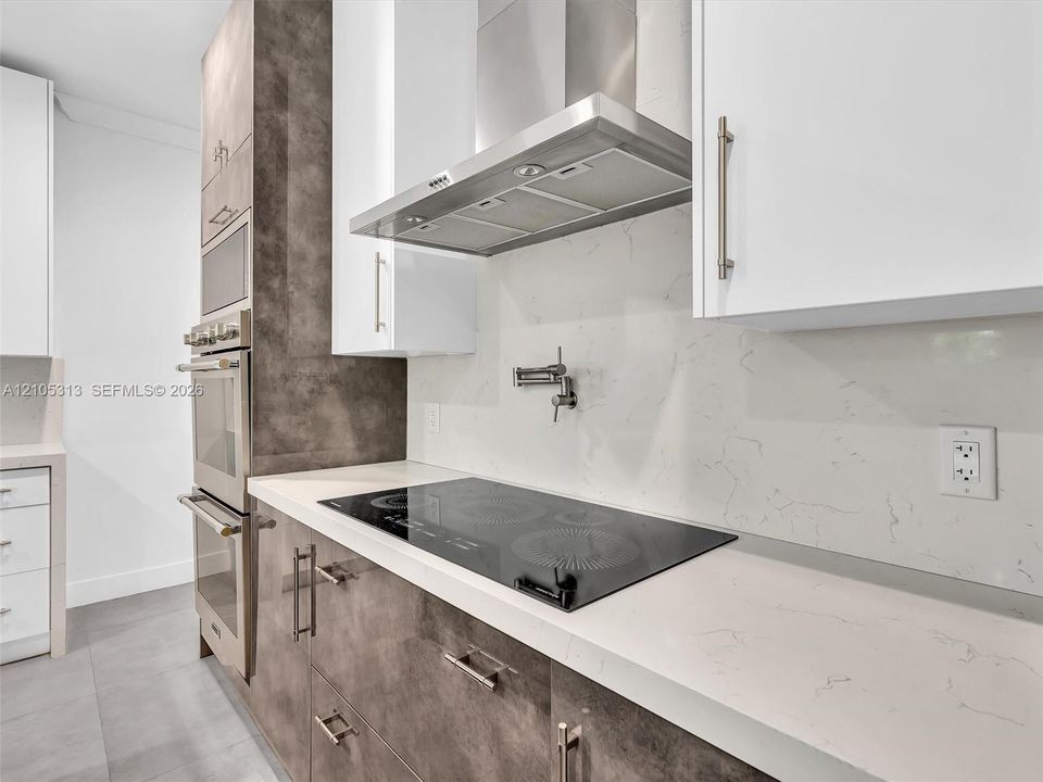 Kitchen featuring white flat-panel cabinetry, contrasting textured brown base cabinetry, a stone-patterned backsplash, a built-in stainless steel double oven, and a cooktop with a wall-mounted pot filler