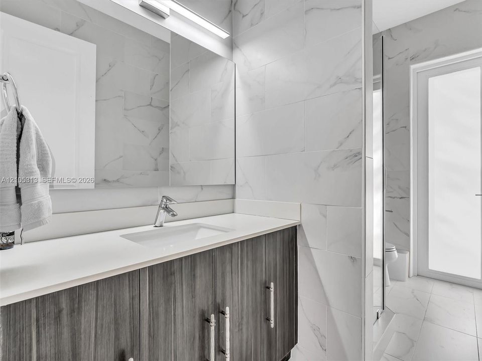 Vanity featuring dark wood-finish cabinetry, a rectangular undermount sink, and a solid-surface countertop, complemented by floor-to-ceiling stone-look wall tile and integrated strip lighting
