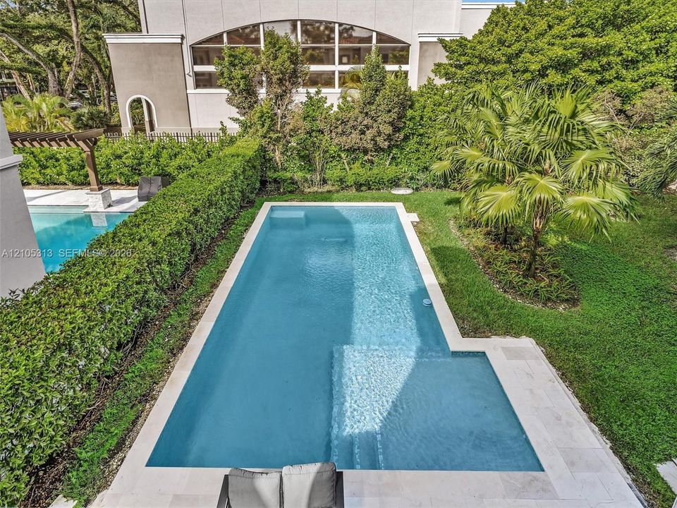 Rectangular swimming pool featuring a sun shelf, light-colored stone paver decking, and natural landscaping perimeter