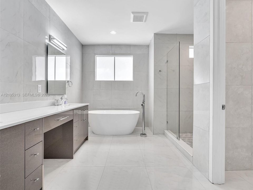 Dual-sink vanity with light-tone countertops, standalone soaking tub, walk-in glass-enclosed shower, and large-format tile flooring