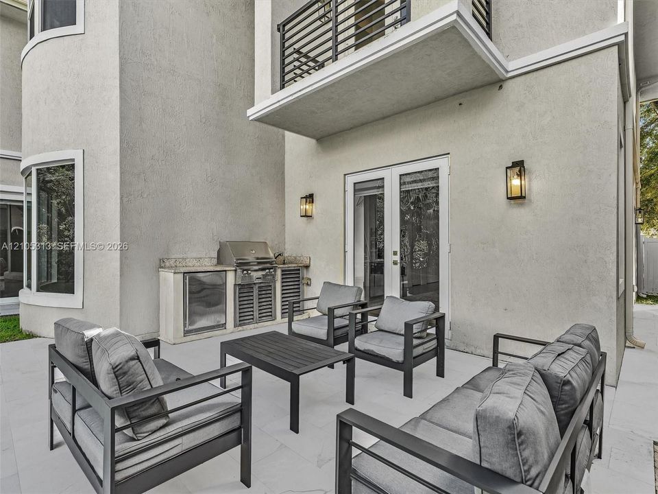 Patio space featuring stone flooring, a built-in outdoor kitchen with stainless steel grill and stone countertop, double glass doors, wall-mounted exterior lanterns, and a second-story balcony with metal railings