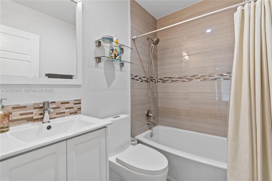 White vanity with integrated sink, chrome faucet, tiled backsplash, white toilet, and a tiled shower/tub combination with a decorative accent band