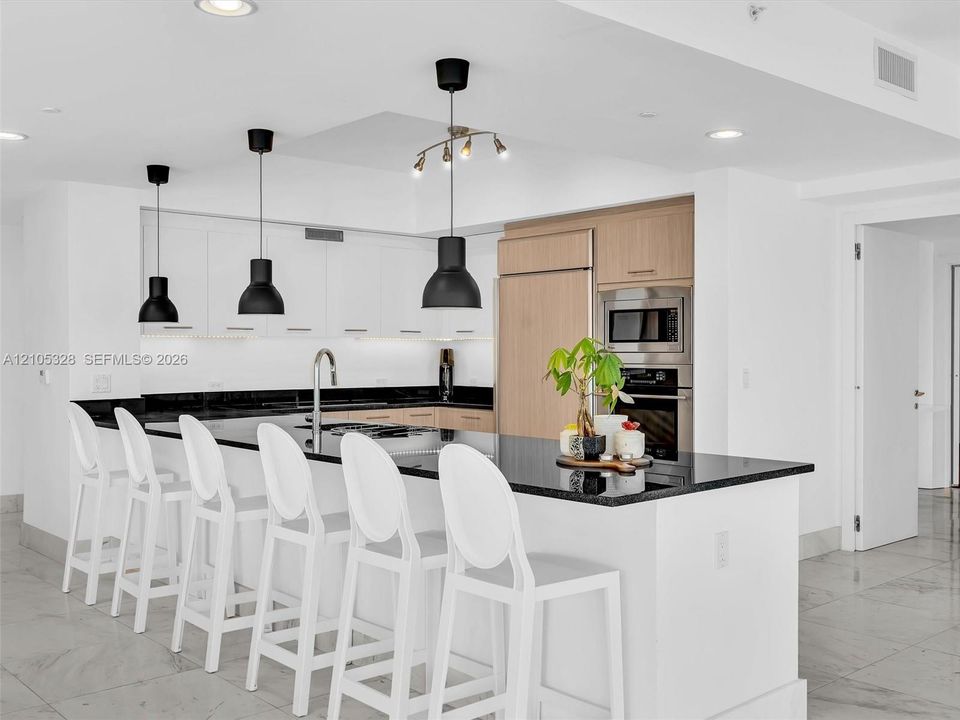 Modern gourmet kitchen featuring an oversized center island with seating, sleek dark stone countertops, stainless steel appliances, and wood-finish cabinetry.