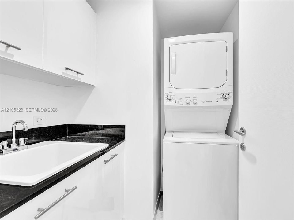 Stacked washer and dryer unit, white cabinetry with silver-tone pulls, black countertop, and an undermount sink with a chrome faucet