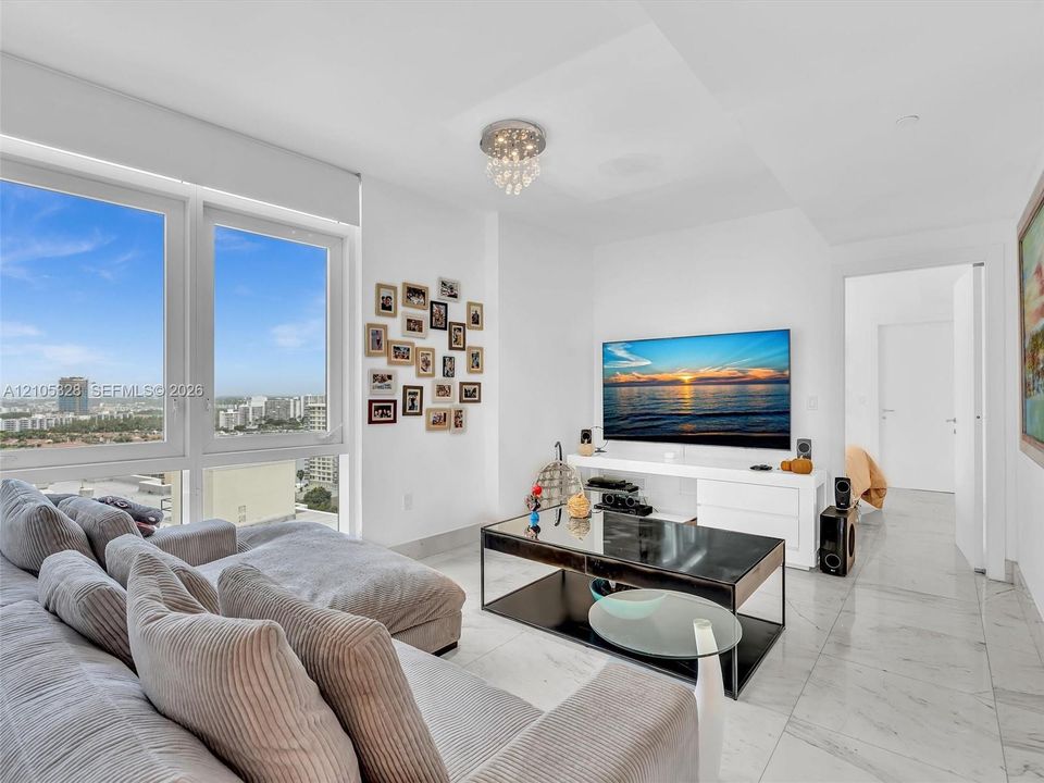 A luxurious and inviting family room, perfect for relaxing or entertaining, featuring expansive windows with captivating city views, elegant white marble-look flooring, recessed lighting, and a stunning crystal chandelier