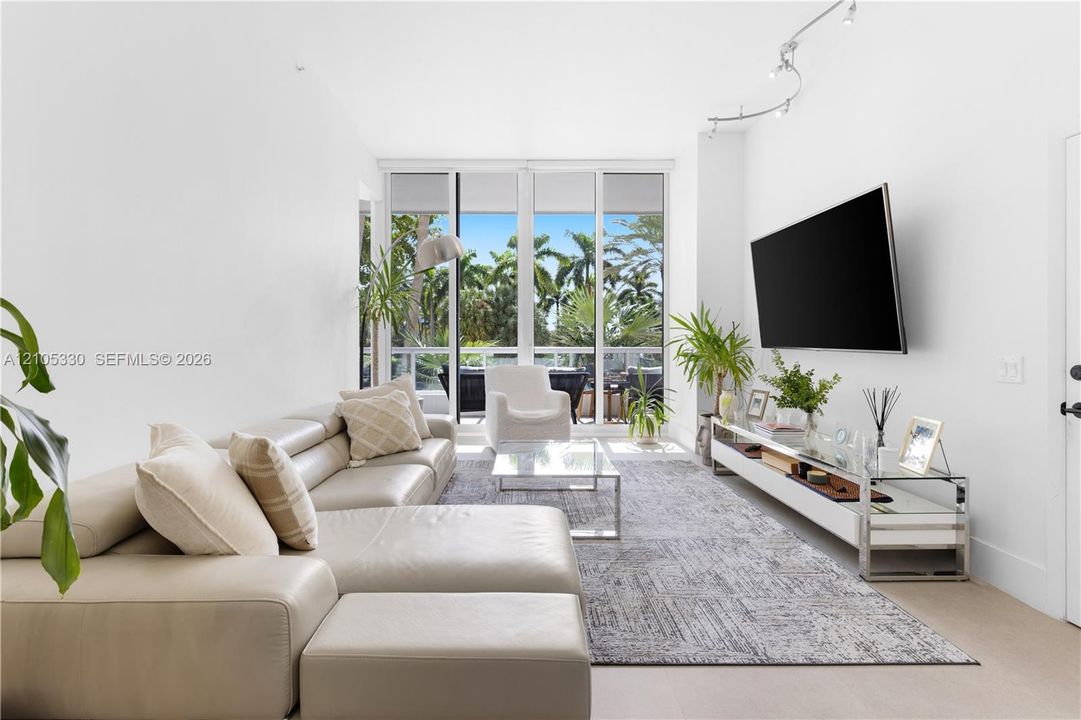 Bright, light-filled living space showcasing floor-to-ceiling glass doors opening to outdoor grounds, neutral wood-finish flooring, and a neutral-toned sectional arrangement