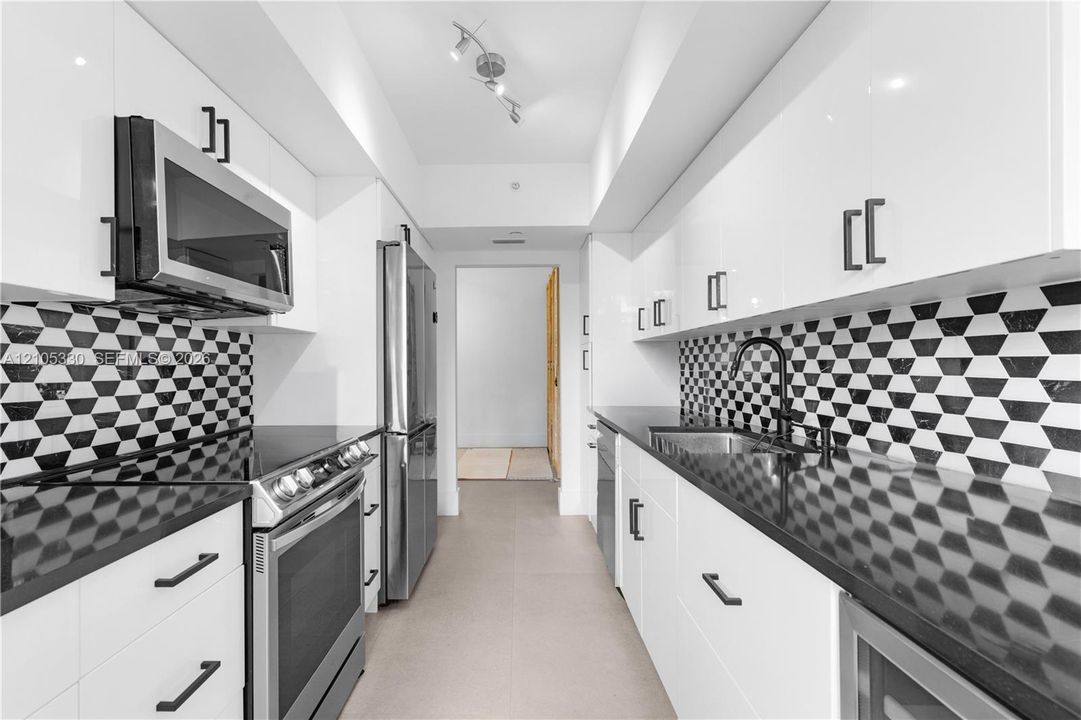 Galley kitchen featuring white cabinetry, stainless steel appliances, black countertops, a geometric pattern backsplash, and track lighting