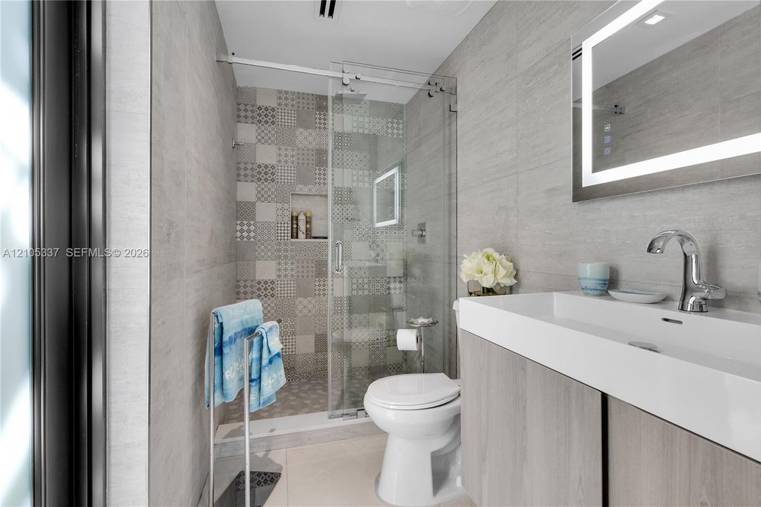 Modern bathroom featuring a wood-finish vanity with an integrated basin, LED-backlit mirror, and a walk-in shower with patterned tile accents, glass enclosure, and a built-in recessed shelf