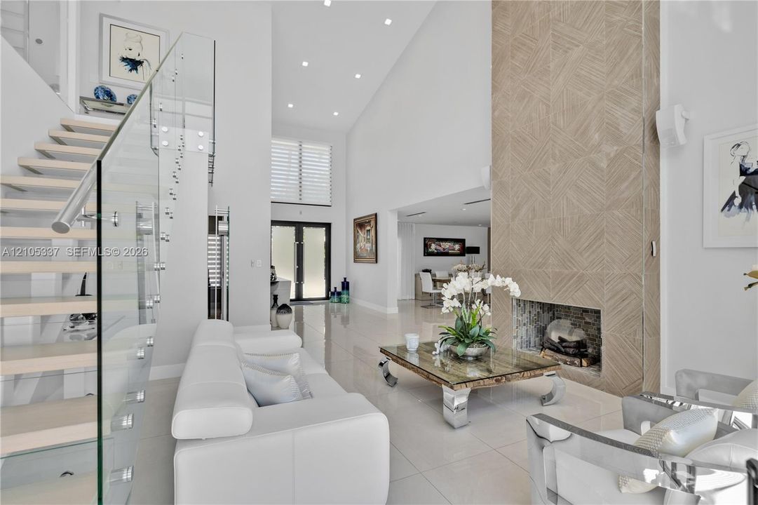 Double-height ceilings, floating staircase with glass panel railing, floor-to-ceiling tiled fireplace surround, and recessed lighting