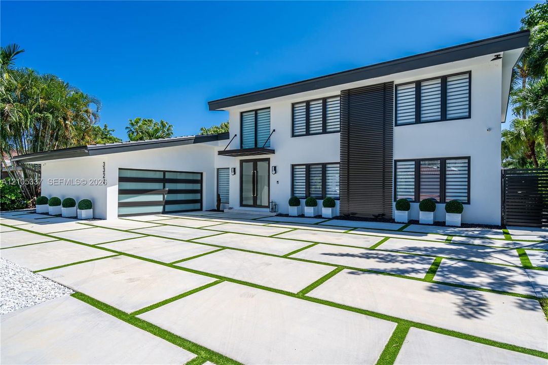 Modern two-story exterior featuring white stucco siding, a horizontal wood-slat accent wall, grid-patterned garage door, and geometric paver driveway with turf infill