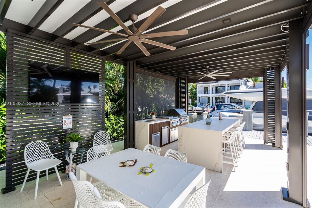 Covered outdoor kitchen featuring a stone-topped island with integrated seating, built-in stainless steel appliances, slatted wood-finish privacy screens, and dual ceiling fans