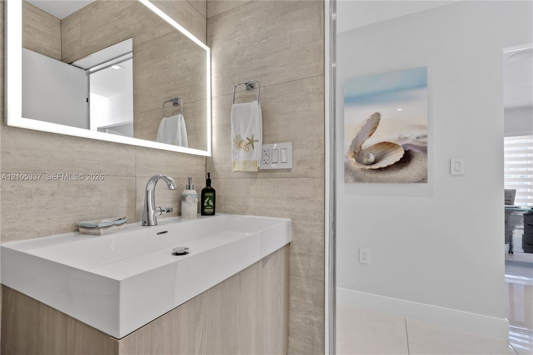 Floating vanity featuring an integrated rectangular sink, wall-mounted polished chrome faucet, and backlit LED mirror, set against large-format neutral wall tile