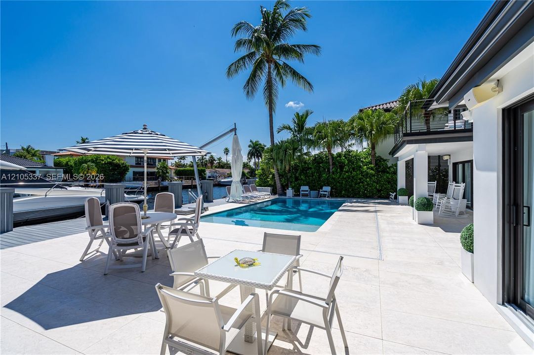 Waterfront patio featuring expansive light-toned stone decking, an integrated in-ground swimming pool, private boat dockage, and a balcony with black metal railing