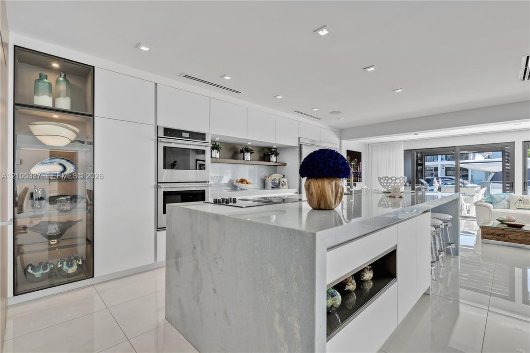 Modern kitchen featuring floor-to-ceiling glass-front cabinetry, a waterfall-edge stone island, built-in dual wall ovens, recessed ceiling lighting, and tile flooring