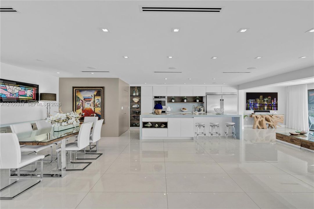 Open-concept kitchen featuring minimalist white cabinetry, a large central island with seating, polished stone-finish flooring, and recessed ceiling lighting