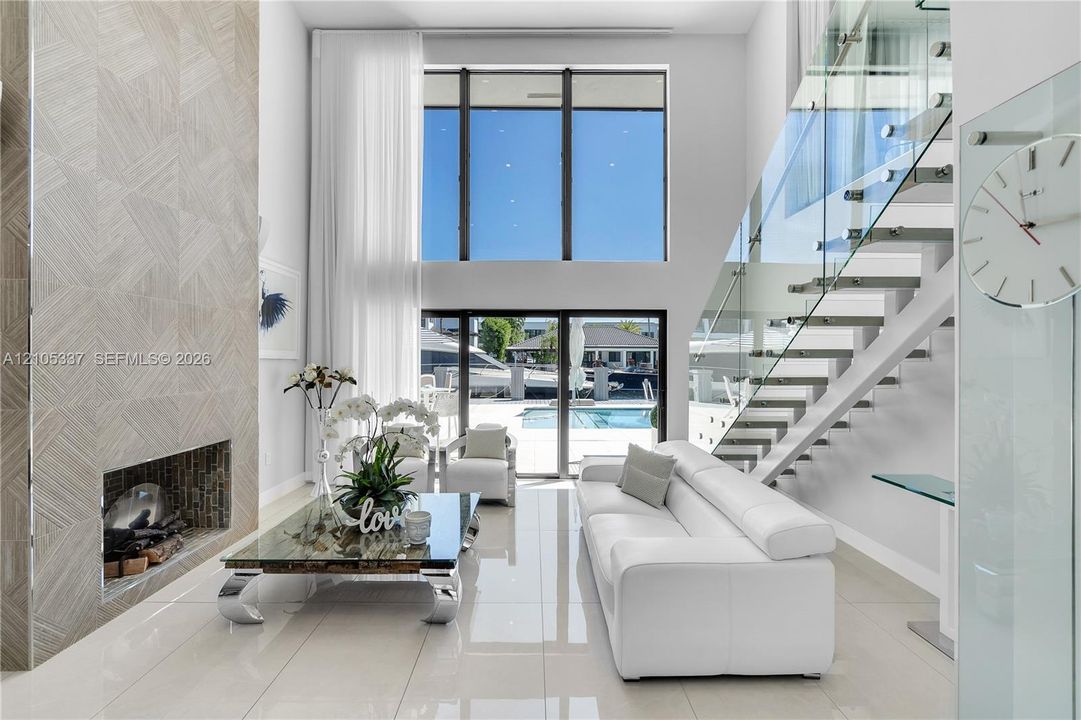 Double-height living area featuring floor-to-ceiling windows, a floor-to-ceiling textured tile fireplace, polished tile flooring, and a staircase with glass railings and metal treads
