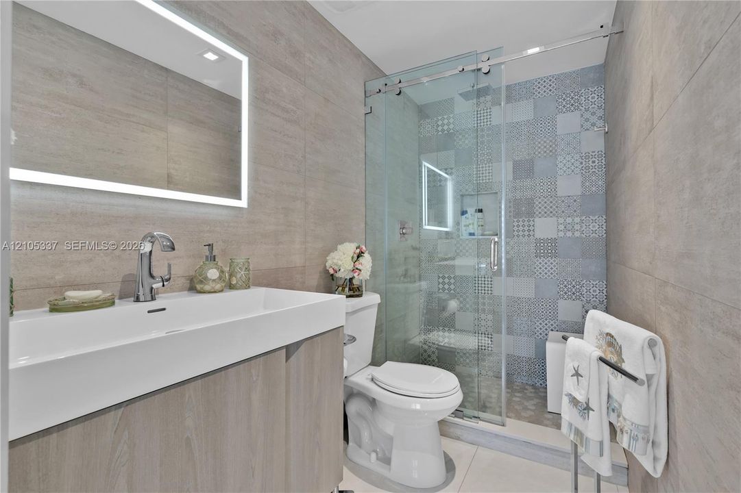 Wall-mounted rectangular basin with a single-handle faucet, integrated LED-backlit mirror, wood-finish vanity cabinetry, glass-enclosed shower with decorative patterned tile wall, and large-format wall tiling