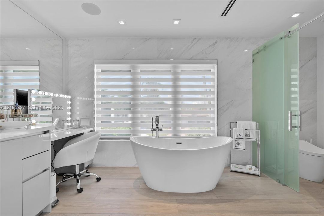 Freestanding soaking tub with a chrome floor-mounted faucet, wall-to-wall stone surfacing, wood-finish flooring, and a frosted glass-enclosed shower