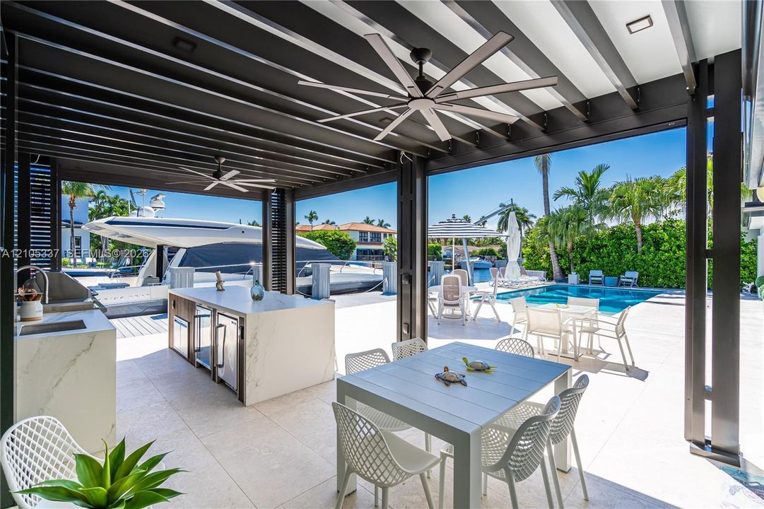 Covered outdoor kitchen featuring a stone-clad island, stainless steel built-in grill, and stone-finish flooring beneath a slatted pergola with multi-blade ceiling fans