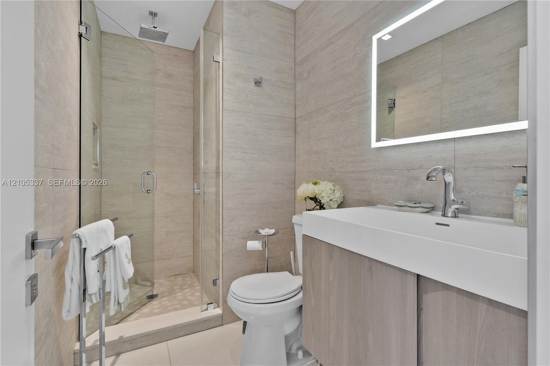 Bathroom featuring large-format neutral wall tile, walk-in glass shower with integrated niche, floating vanity with vessel-style sink, and LED-backlit mirror