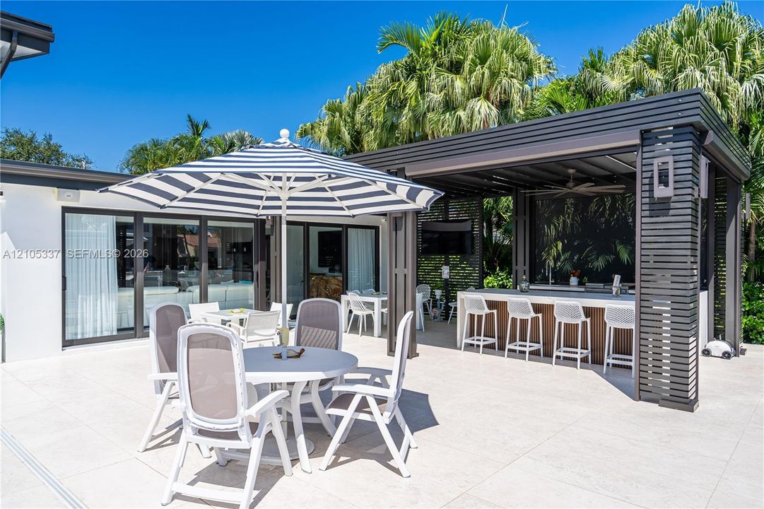 Outdoor patio featuring large-format light-toned tiling, a covered cabana with a built-in stone-top bar, recessed ceiling fan, and sliding glass doors