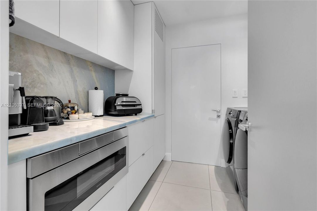 Utility room featuring flat-panel white cabinetry, stone-surface countertop, full-height stone backsplash, built-in stainless steel microwave, and large-format floor tile