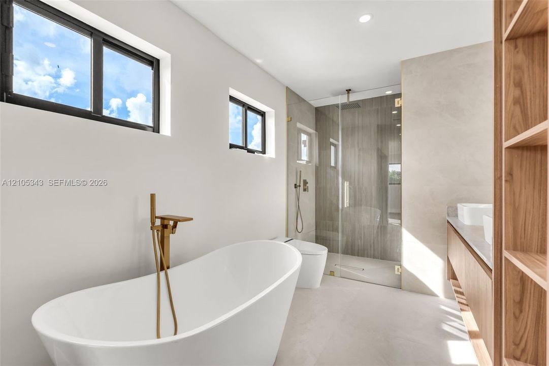 Freestanding soaking tub with a brass floor-mounted faucet, walk-in glass-enclosed shower, wall-mounted dual-flush toilet, floating wood-finish vanity with countertop sinks, and built-in wood shelving