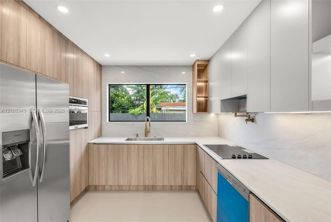Modern kitchen featuring flat-panel wood-finish cabinetry, white stone-composite countertops, stainless steel appliances, recessed ceiling lighting, and a wall-mounted pot filler faucet