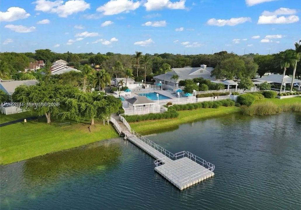 Waterfront property featuring a private dock, expansive green lawn, mature trees, and a community pool area with a covered patio