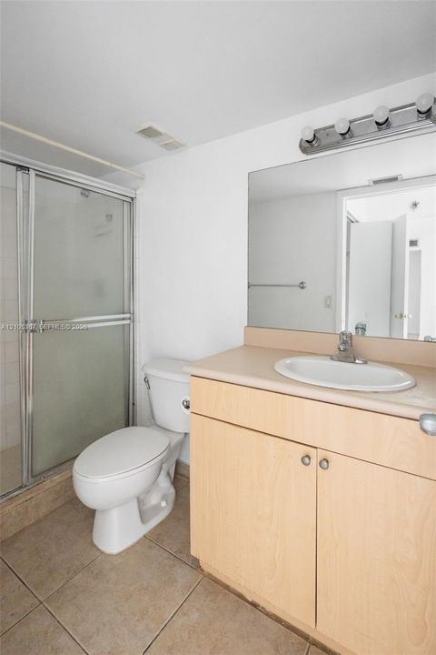 Bathroom featuring light wood-finish cabinetry, a neutral countertop with an integrated oval basin, ceramic floor tiling, a walk-in shower with a glass enclosure, and a wall-mounted four-bulb vanity fixture