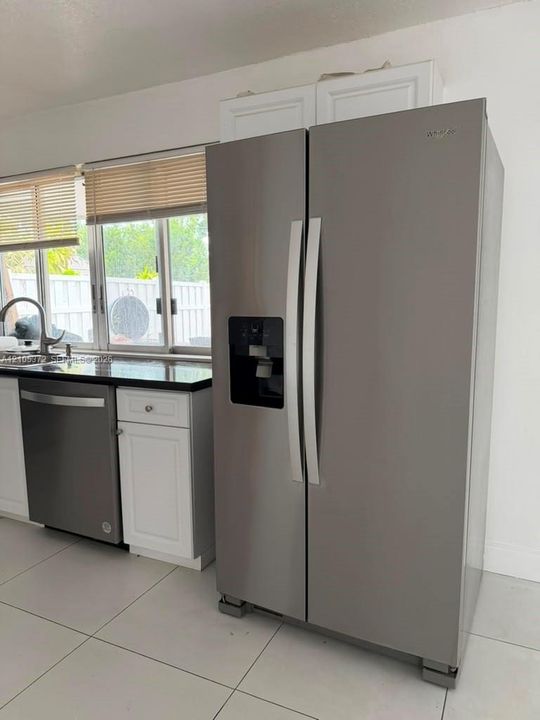 Stainless steel side-by-side refrigerator, stainless steel dishwasher, white cabinetry, dark stone countertop, and tile flooring