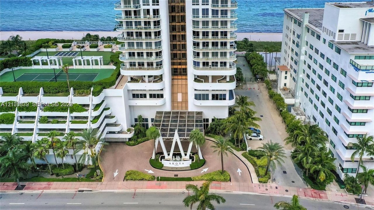 High-rise beachfront property featuring a multi-story glass facade, tennis court, porte-cochère entry with a central fountain, and surrounding mature landscaping