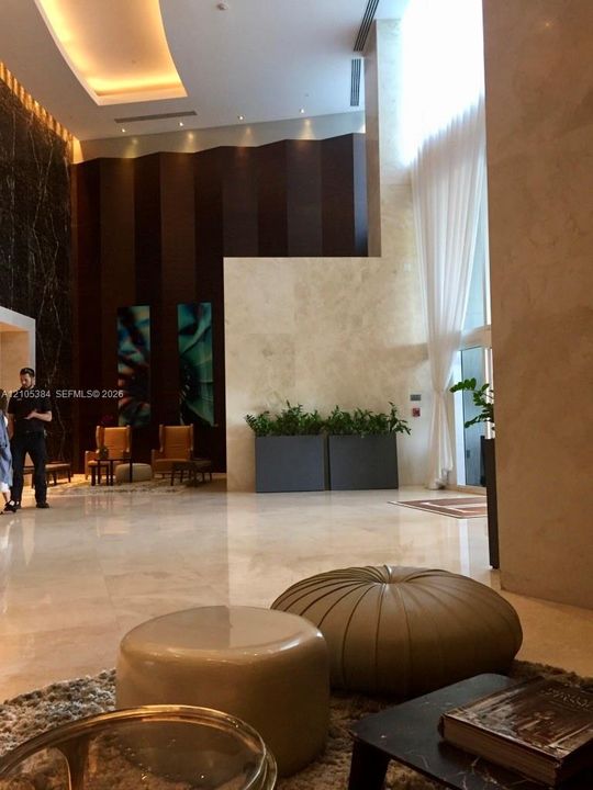 High ceilings with recessed lighting, full-height window with sheer drapes, dark wood-paneled accent wall, light-toned stone flooring, and built-in planters