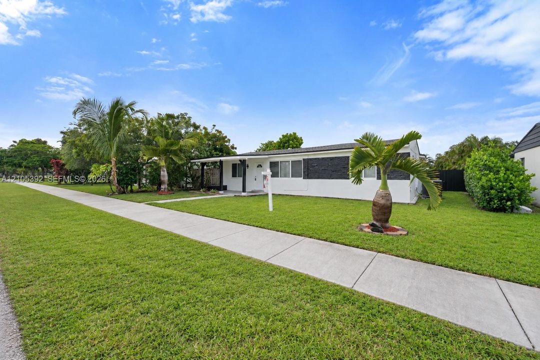 White exterior with black accent siding, covered entry, front lawn, and mature palm trees