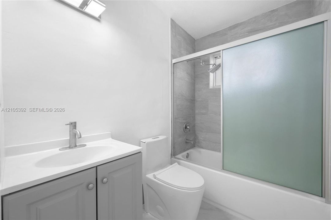 Bathroom featuring a vanity with a white countertop and gray cabinetry, a toilet, a tiled shower/tub combination with a sliding frosted glass enclosure, and a chrome faucet