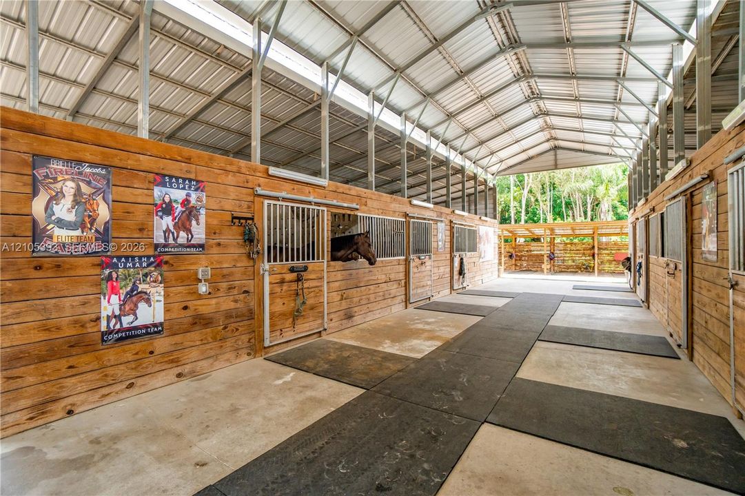 Equestrian facility featuring wood-paneled walls, individual metal-framed stalls, a high-clearance metal roof with translucent skylight panels, and rubber-mat flooring