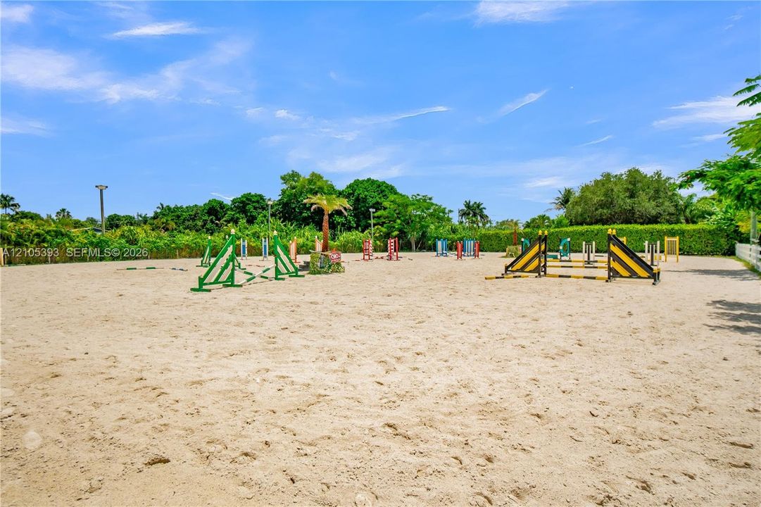 Sand equestrian arena featuring professional-grade jumping obstacles, white perimeter fencing, and mature surrounding landscaping