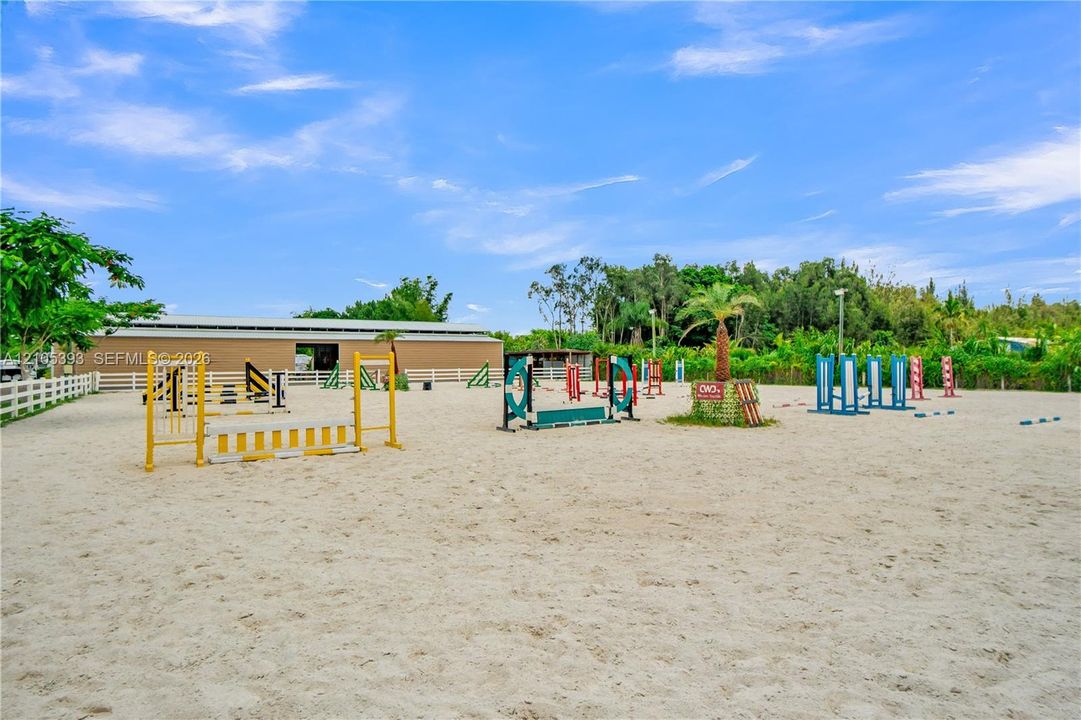Equestrian arena featuring a sand surface, multiple jump obstacles, perimeter white rail fencing, and a detached structure