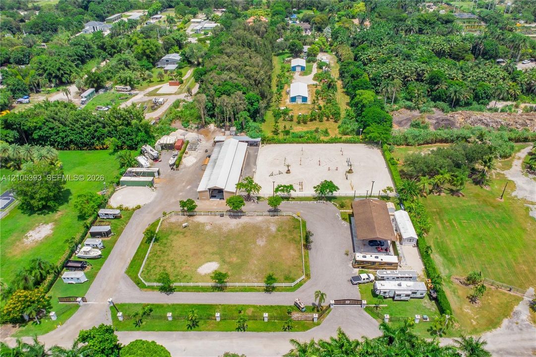 Aerial perspective of an expansive equestrian facility featuring a fenced riding arena, a long stable structure with a metal roof, dedicated paddocks, and paved driveways