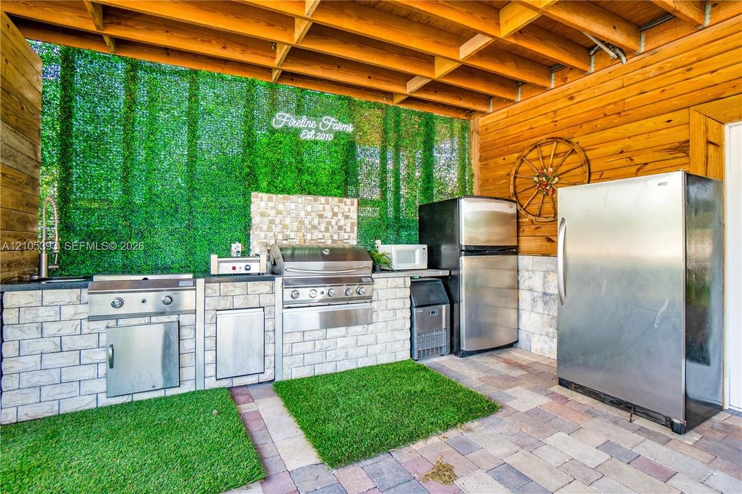 Covered outdoor kitchen featuring light-toned brick masonry, stainless steel built-in grill, stone tile backsplash, integrated refrigeration, and wood-paneled ceiling