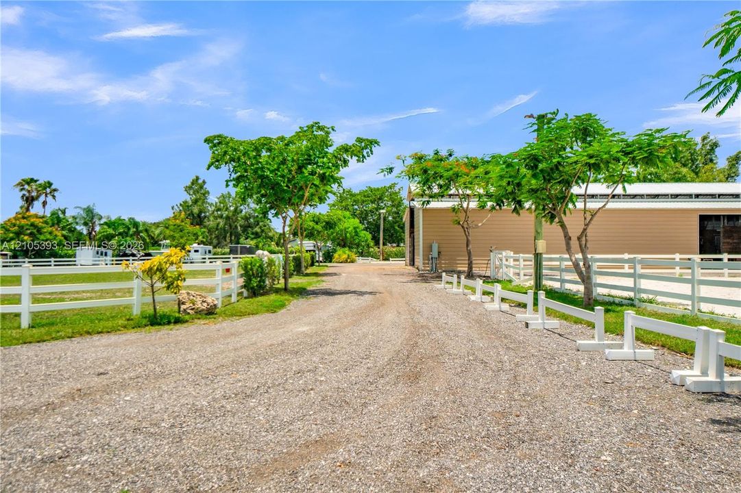 Gravel driveway leading to a metal-sided barn, featuring white split-rail fencing, mature trees, and open grounds