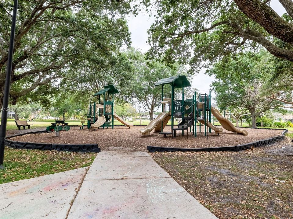 Outdoor recreational area featuring multiple play structures, a concrete walking path, and mature tree canopy