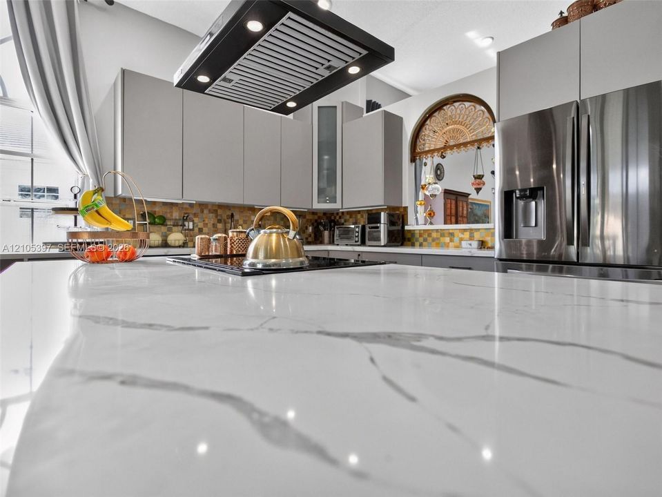 Kitchen featuring a large waterfall-edge island with stone-finish countertop, flat-panel cabinetry, stainless steel refrigerator, and built-in rectangular range hood