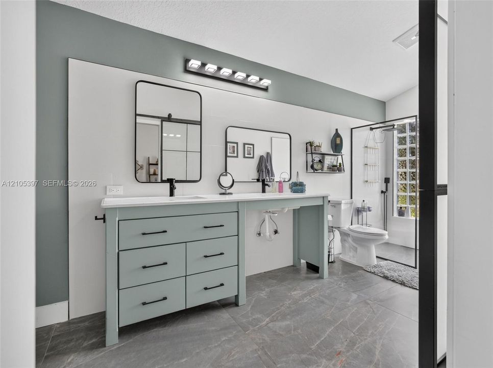 Dual-sink vanity with matte black hardware, rectangular wall-mounted mirrors, stone-finish tile flooring, integrated light fixture, and glass-enclosed shower stall ADA accessible