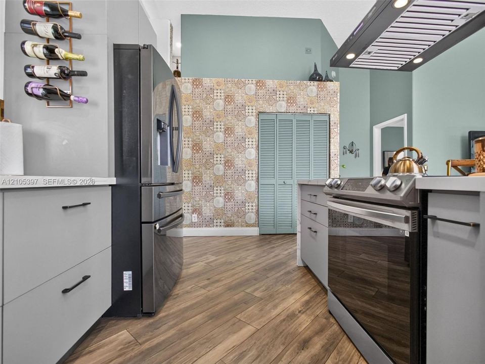 Kitchen featuring grey flat-panel cabinetry, wood-finish flooring, patterned tile accent wall, stainless steel appliances, and a mounted range hood