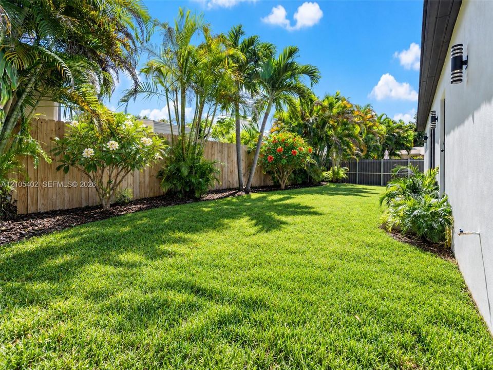 Landscaped backyard featuring a manicured lawn, wood-panel fencing, mature palm trees, and exterior wall-mounted light fixtures