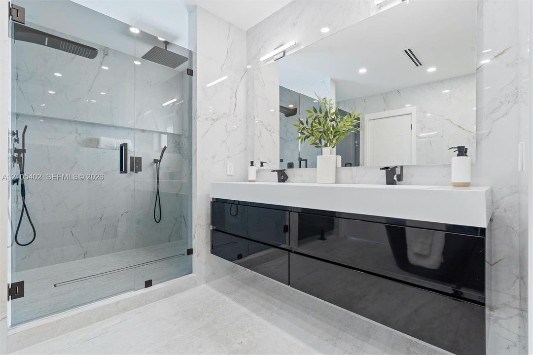 Floating high-gloss black vanity featuring a dual-sink white countertop, wall-to-wall mirror, glass-enclosed walk-in shower with stone-finish wall tile, and matte black fixtures