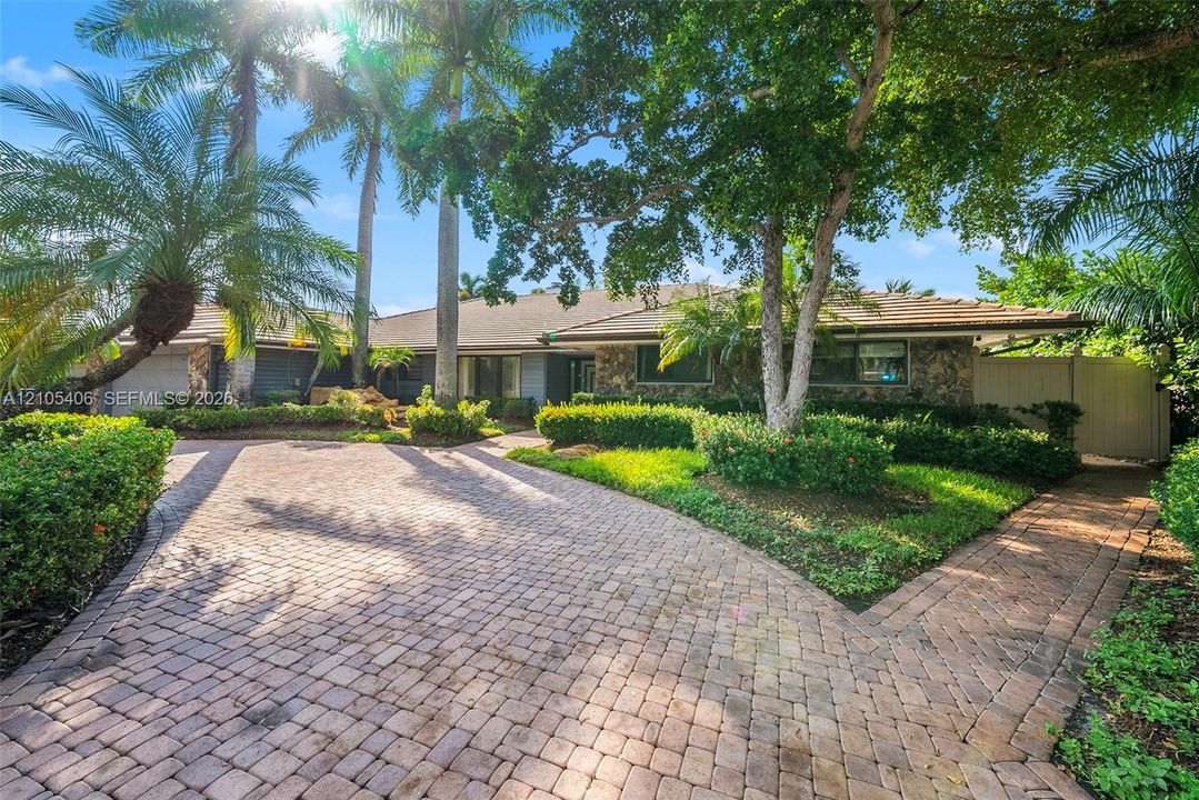 Stone-clad exterior with a tiled roof, paver driveway, and lush landscaping, featuring mature palm trees and established hedging